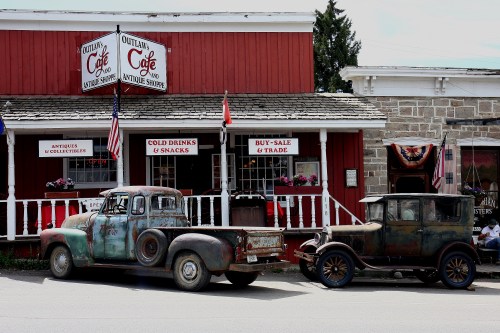 Two vintage, weathered trucks parked in front of a red café with a porch, flags, and signs for drinks and antiques.