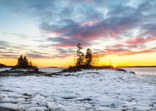 A serene winter landscape featuring ice-covered rocks and trees at sunset, with colorful clouds in the sky.