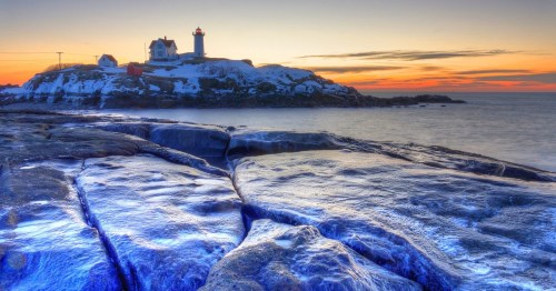 A lighthouse on a rocky shore at sunset, with snow and a colorful sky reflecting on the water.