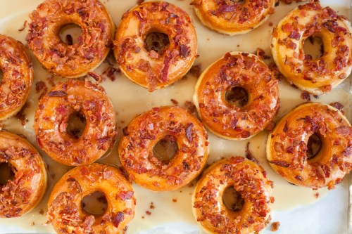 A tray of glazed donuts topped with crispy bacon bits, arranged neatly on parchment paper.