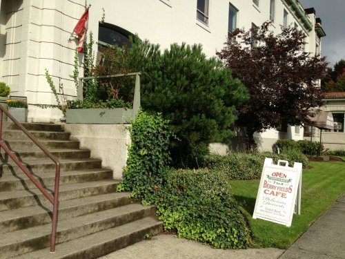 A cafe sign outside a building with stairs, greenery, and a flag, indicating the cafe is open for business.