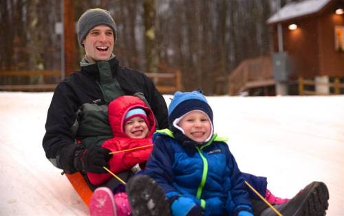 A man and two children smile while sledding down a snowy hill, surrounded by trees and a cozy cabin in the background.