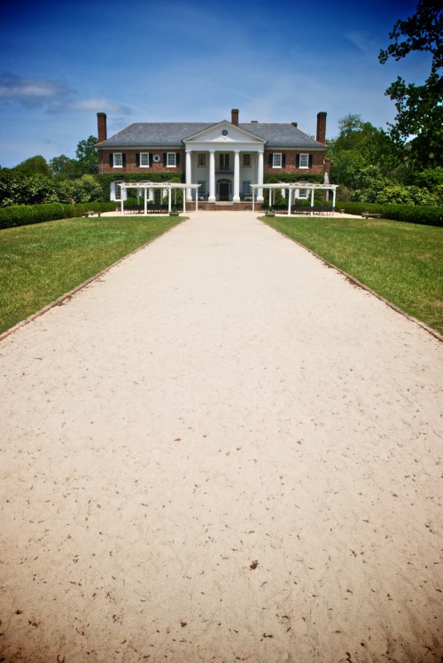 A grand mansion with a white facade, flanked by green lawns and a sandy pathway leading to the entrance.