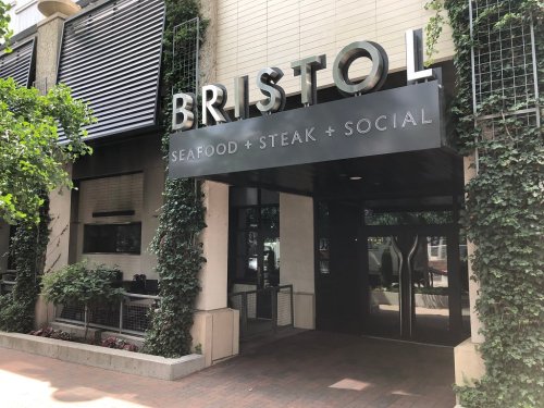 Entrance of Bristol restaurant featuring a sign that reads "Seafood + Steak + Social," surrounded by greenery.