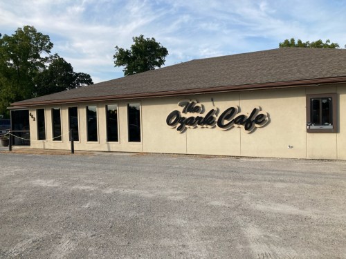 Exterior of The Ozark Café, featuring a beige building with large windows and a sign on the wall. Gravel parking area.