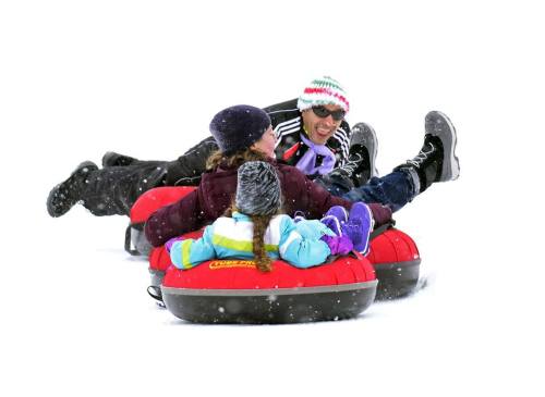 Three people enjoy tubing down a snowy hill, laughing and having fun in winter attire. Snowflakes are falling around them.