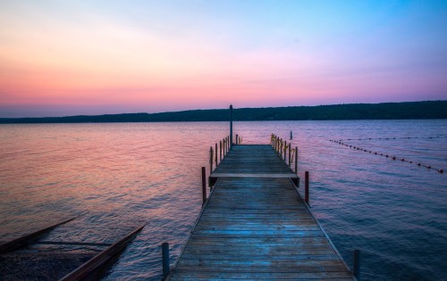 A serene wooden dock extends into a calm lake at sunset, with soft pink and blue hues in the sky.