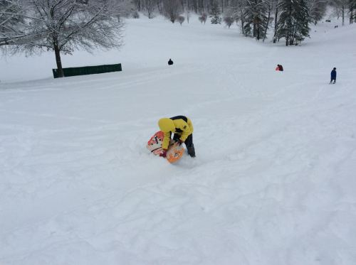 A person in a yellow jacket struggles to stand while holding a sled on a snowy hill, with others playing in the background.