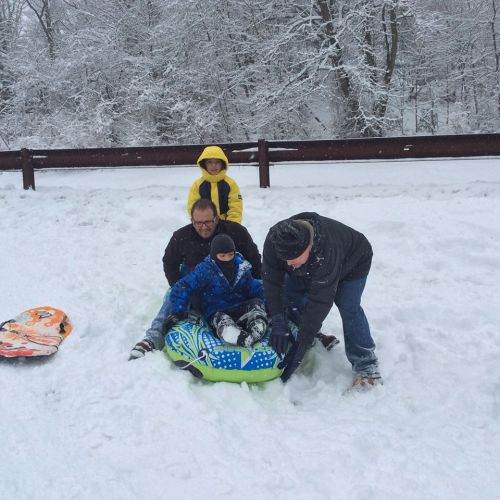 A snowy scene with four people, two on sleds, enjoying winter fun in a snowy landscape. Trees are covered in snow.