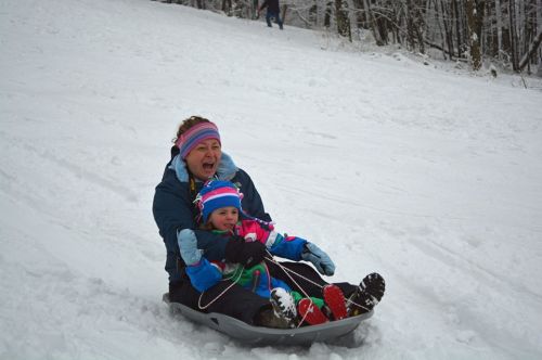 A woman and a child joyfully sledding down a snowy hill, both wearing colorful winter clothing.