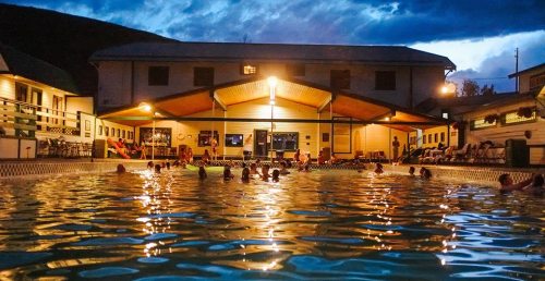 A twilight scene of a pool with people swimming, surrounded by a building and mountains under a cloudy sky.