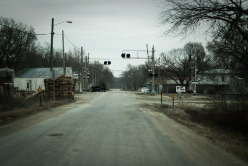 A quiet, empty road with a railroad crossing ahead, surrounded by bare trees and sparse buildings.