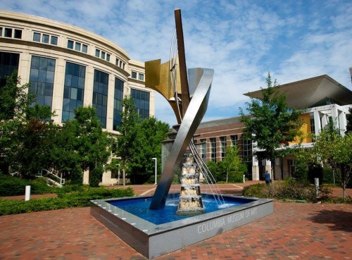 Sculpture with water feature in front of the Columbia Museum of Art, surrounded by greenery and blue sky.