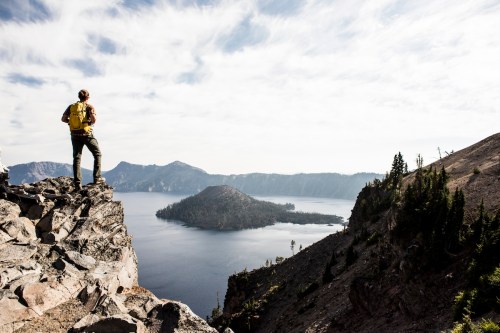 A hiker stands on a rocky ledge overlooking a serene lake surrounded by mountains and trees under a cloudy sky.