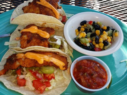 Three fish tacos with toppings on a plate, accompanied by a bowl of black bean salad and a cup of salsa.