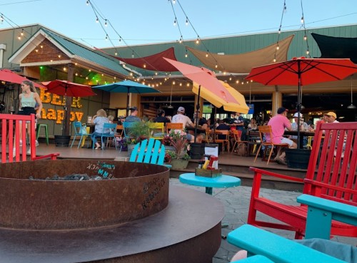 Outdoor dining area with colorful umbrellas, string lights, and a fire pit, filled with people enjoying a lively atmosphere.
