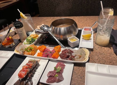 A table set with a variety of meats, vegetables, and sauces for a fondue meal, alongside drinks and garnishes.