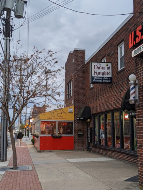 A street view featuring brick buildings, a food stand, and a tree with bare branches under a cloudy sky.