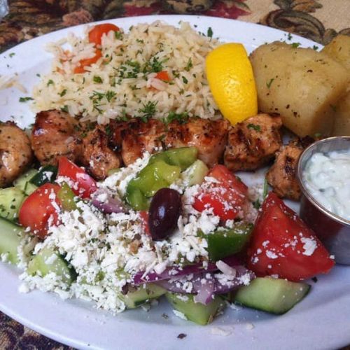A plate of grilled chicken skewers, rice, potatoes, and a fresh salad with feta cheese and a side of tzatziki sauce.