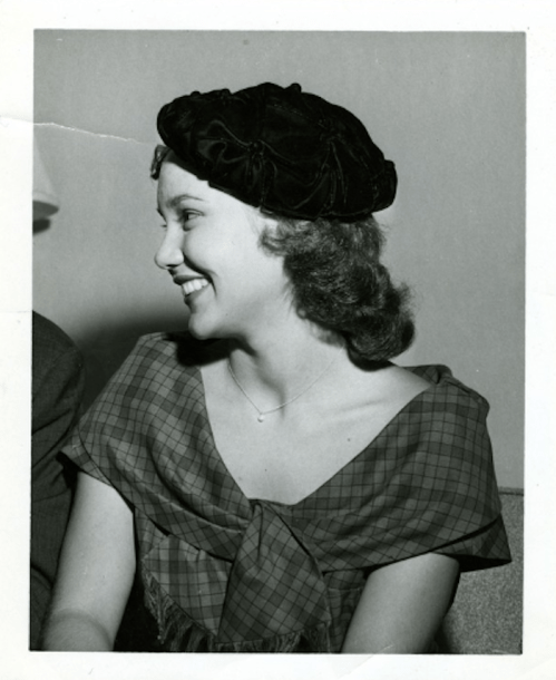 A smiling woman in a vintage dress and black hat, captured in profile against a neutral background.