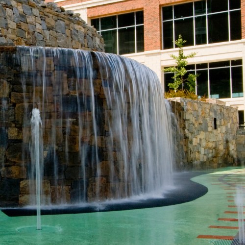 A cascading waterfall flows over a stone structure into a circular pool, surrounded by greenery and modern buildings.