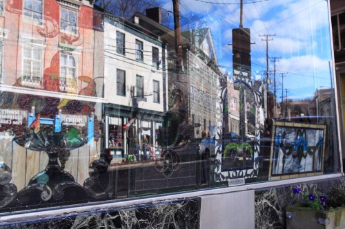 A reflective window showcasing colorful designs and the street scene behind it, with buildings and power lines visible.