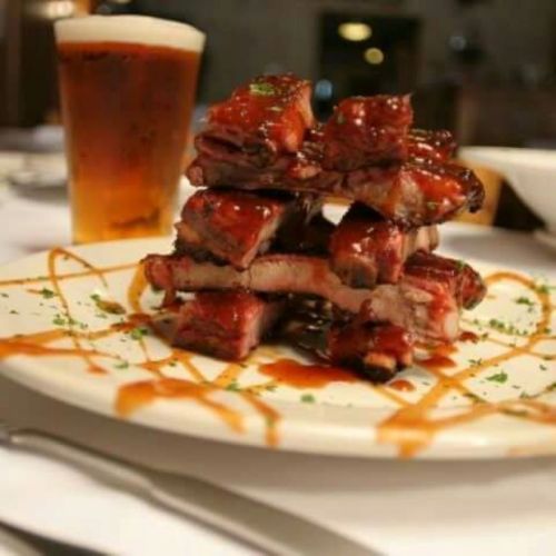 A stack of glazed ribs on a plate, garnished with sauce and herbs, alongside a glass of beer in the background.