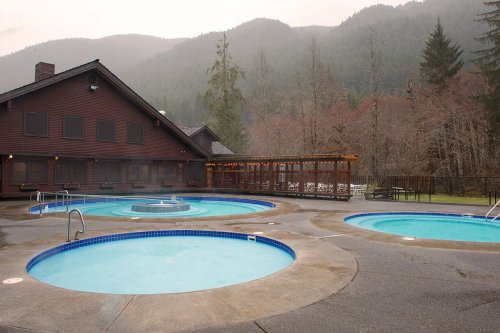 A serene outdoor spa area with multiple hot tubs, surrounded by trees and mountains in a misty atmosphere.