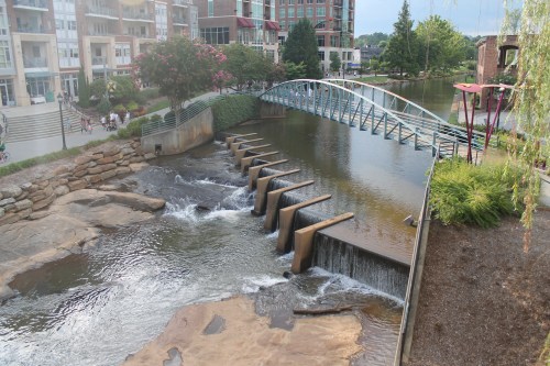 A serene river scene featuring a small dam, a modern bridge, and lush greenery along the water's edge.