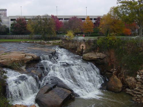 A waterfall cascades over rocky terrain, surrounded by trees with autumn foliage and a parking structure in the background.