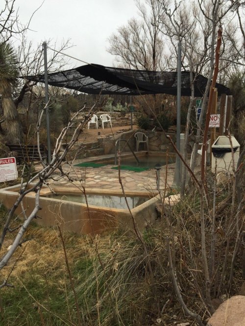 A secluded outdoor area with a hot tub, surrounded by sparse vegetation and a shaded canopy. Signs are visible nearby.