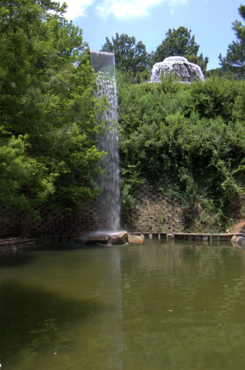 A serene waterfall cascades into a calm pond, surrounded by lush greenery and trees under a clear blue sky.
