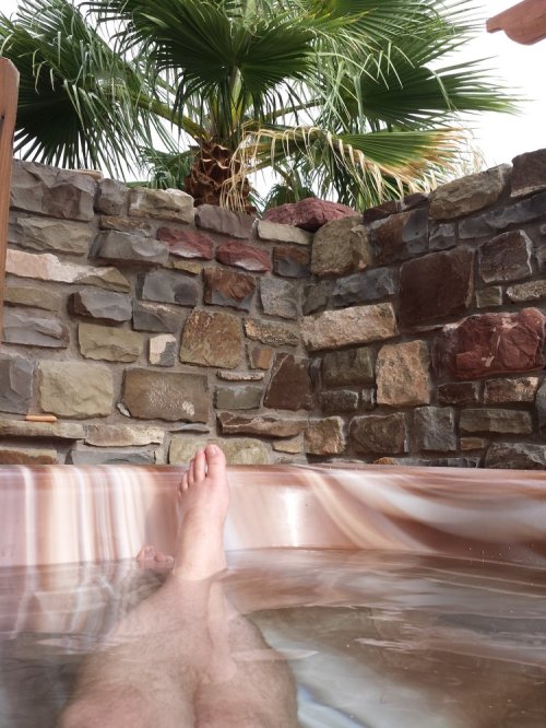 A person's foot resting in a hot tub, with a stone wall and palm trees in the background.