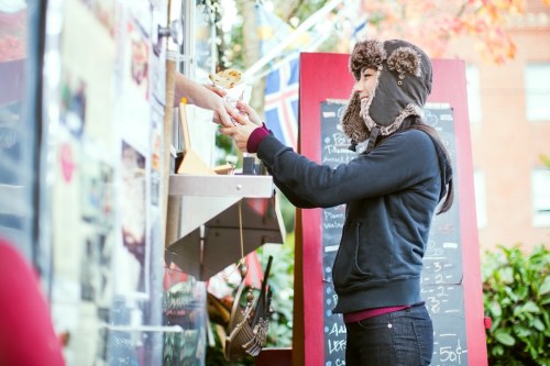 A woman in a winter hat receives food from a food truck, surrounded by colorful decorations and a menu board.