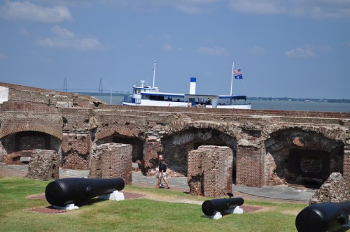A historic fort with brick ruins and large cannons, overlooking a boat on the water under a clear blue sky.