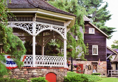 A charming gazebo with intricate white latticework beside a rustic wooden building, surrounded by greenery and flowers.