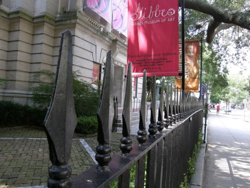 A view of the Gibbs Museum of Art's fence, featuring decorative iron spikes and banners along the sidewalk.