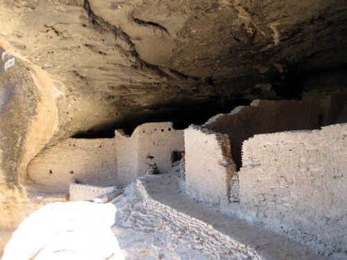 Ancient stone ruins under a rocky overhang, featuring walls and pathways in a desert landscape.