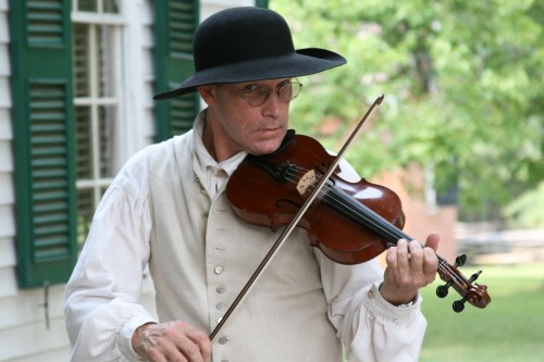 A man in period clothing plays the violin outdoors, wearing a wide-brimmed hat and glasses, with green shutters in the background.
