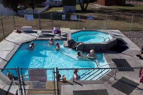 Aerial view of a fenced pool area with people swimming and relaxing in two hot tubs on a sunny day.