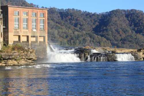 A historic building beside a waterfall, with flowing water and lush green hills in the background under a clear blue sky.