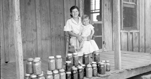 A woman and a child sit on a porch surrounded by jars of preserved food.