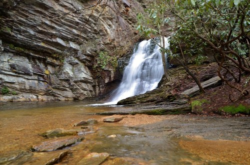 A serene waterfall cascades over rocky cliffs into a calm pool, surrounded by lush greenery and smooth stones.