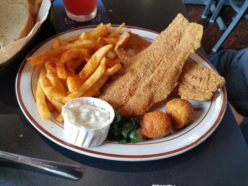 A plate of fried fish fillets, French fries, cornbread balls, and a small cup of tartar sauce on a table.