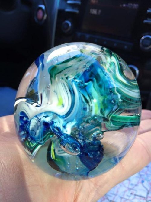 A hand holding a colorful glass marble with swirling blue and green patterns, set against a car interior background.