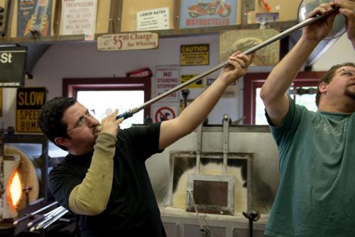 Two men work together in a glassblowing studio, one shaping glass with a long rod while the other assists.