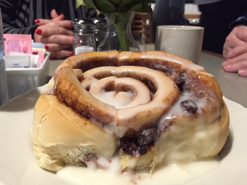 A large, gooey cinnamon roll with cream icing, served on a plate with a coffee cup and sugar packets in the background.