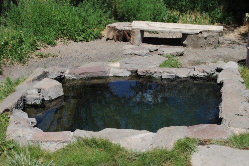 A natural hot spring surrounded by stone, with a wooden bench nearby and greenery in the background.