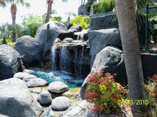 A serene waterfall cascading over rocks, surrounded by lush greenery and colorful plants under a clear blue sky.