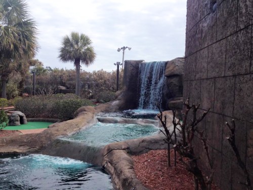 A serene mini-golf scene featuring a waterfall, palm trees, and a winding water feature amidst lush greenery.